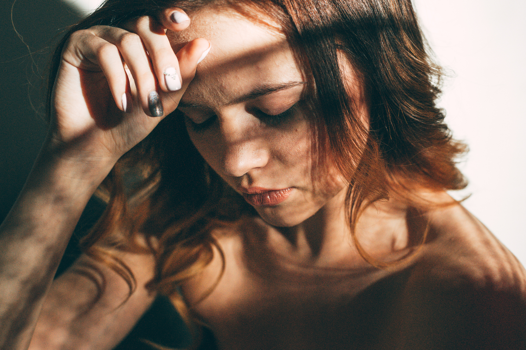 A person with long hair looks down thoughtfully, with their hand resting near their forehead, shadows cast across their face