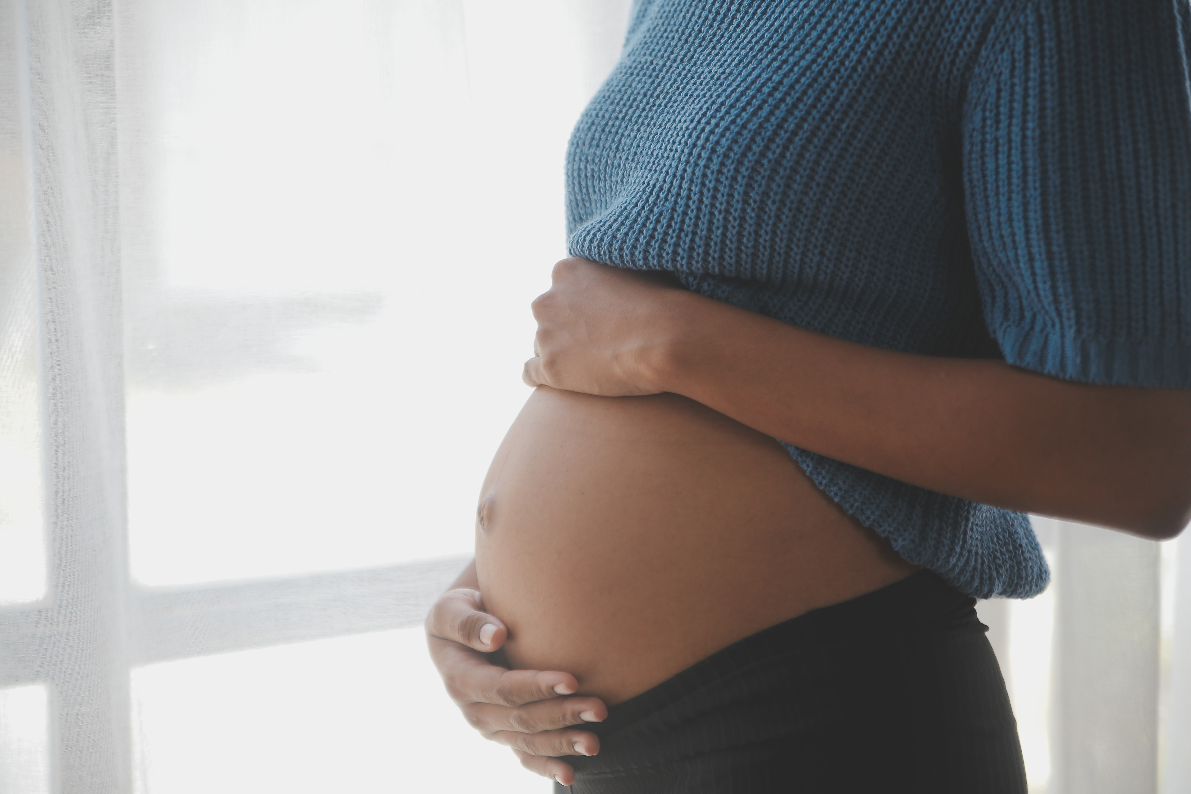 Pregnant person cradling their belly, standing by a window. Focus on connection and anticipation