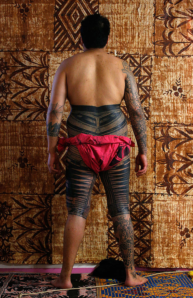 Individual with traditional tattoos stands with back to the camera, wearing a red cloth around the waist