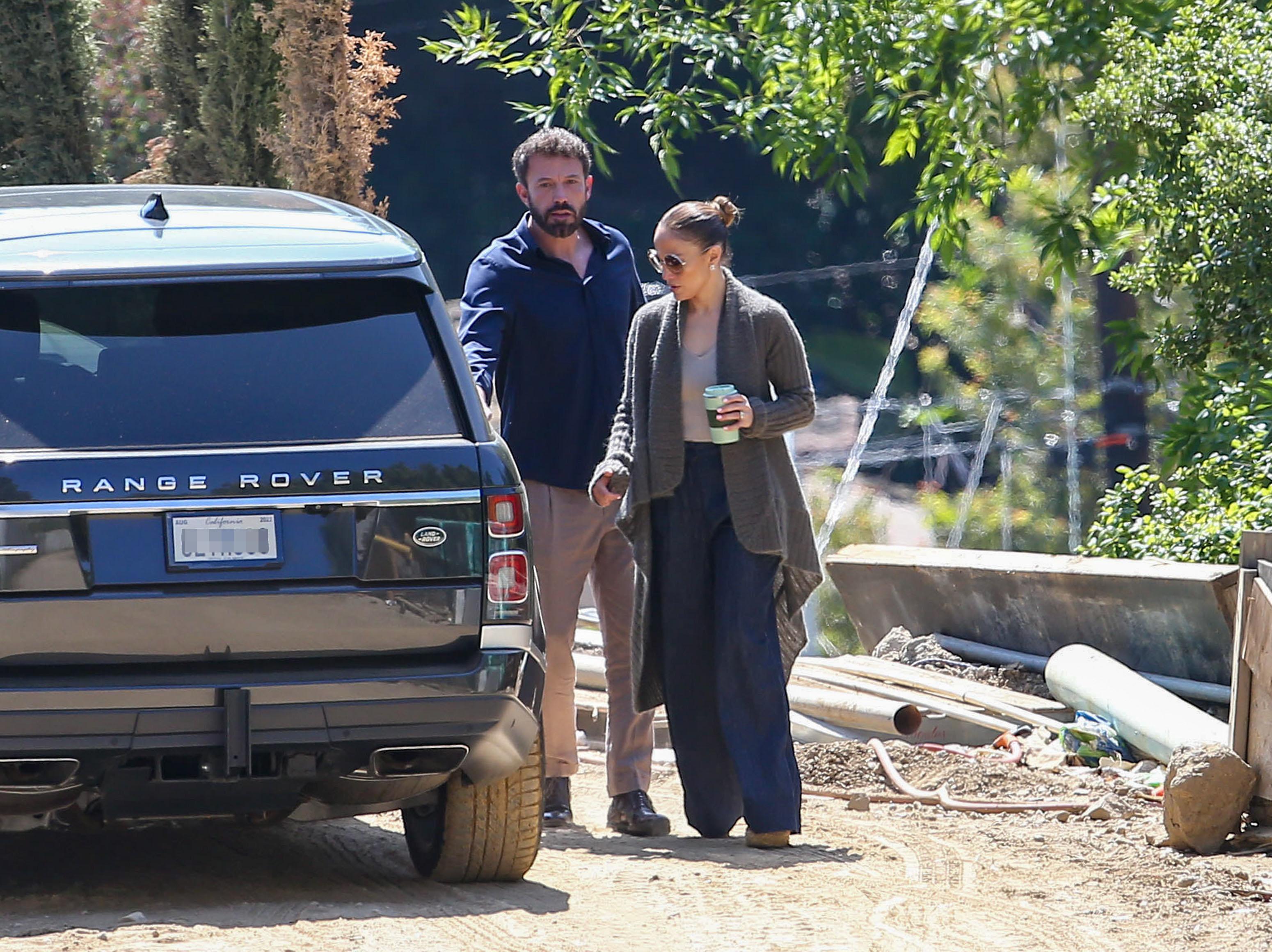 Ben Affleck and Jennifer Lopez casually walk next to a parked Range Rover, with Jennifer holding a drink and wearing a long cardigan and wide-legged pants