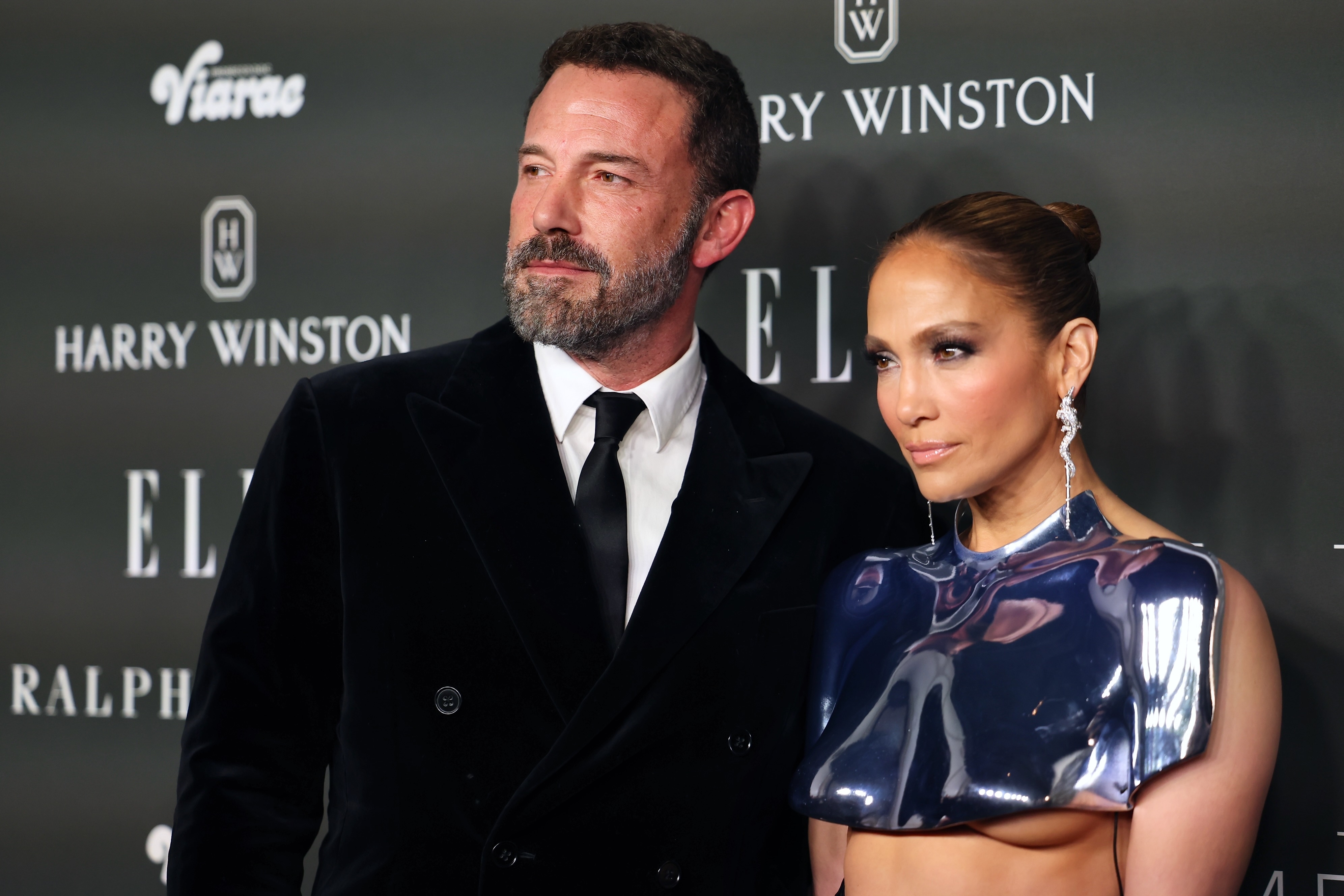 Ben Affleck and Jennifer Lopez pose on the red carpet. Ben wears a black velvet suit and tie; Jennifer wears a futuristic, metallic top with a sleek high bun