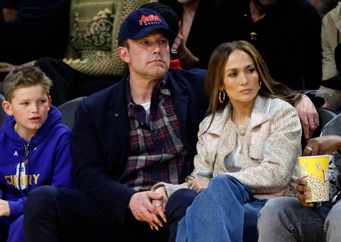 Ben Affleck, Jennifer Lopez, and a young boy sit courtside at a basketball game. Ben wears casual attire with a baseball cap, and Jennifer wears stylish, casual clothing