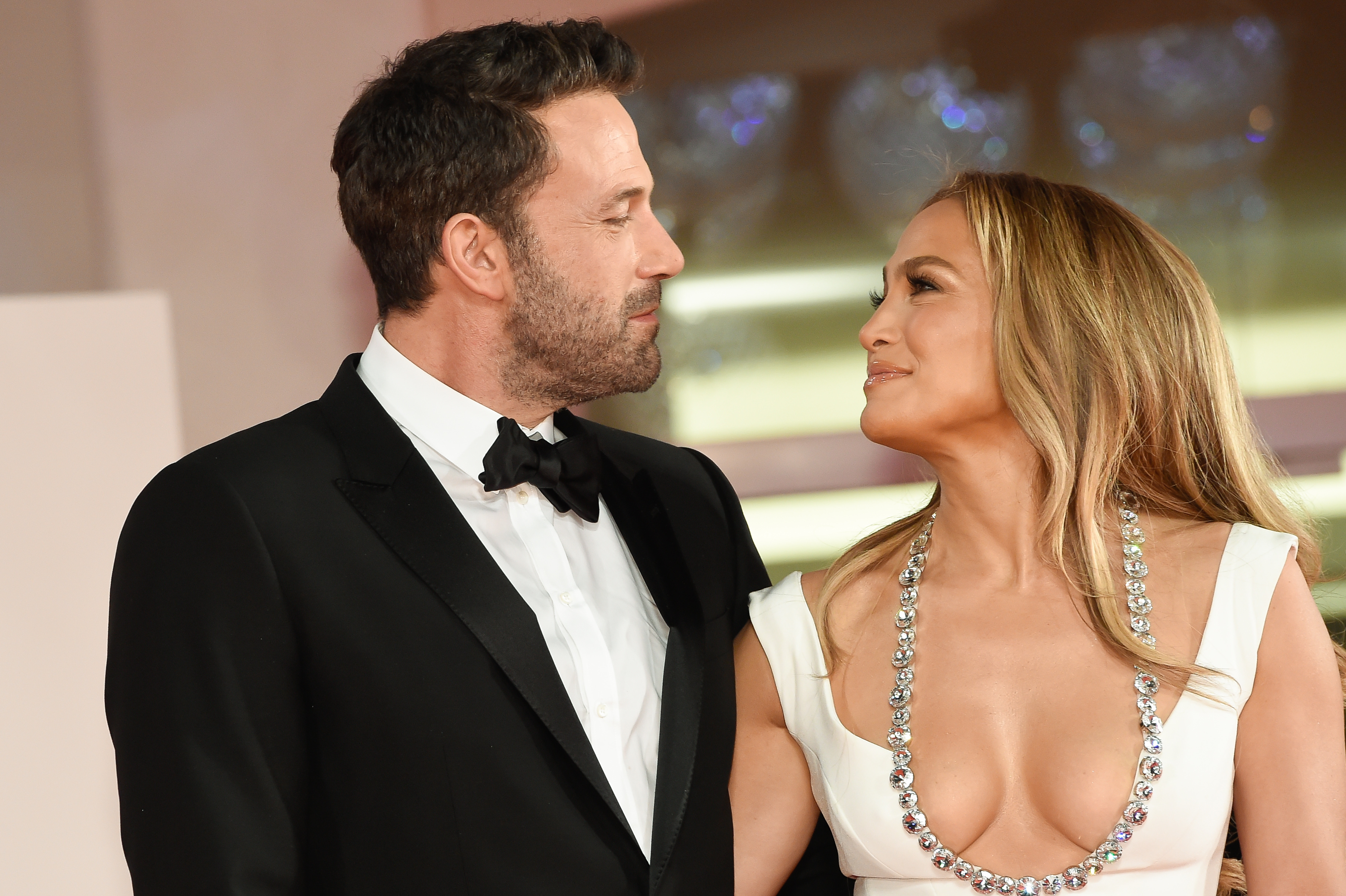 Ben Affleck in a tuxedo and Jennifer Lopez in a low-cut white dress with a sparkling necklace, smiling at each other on a formal red carpet event