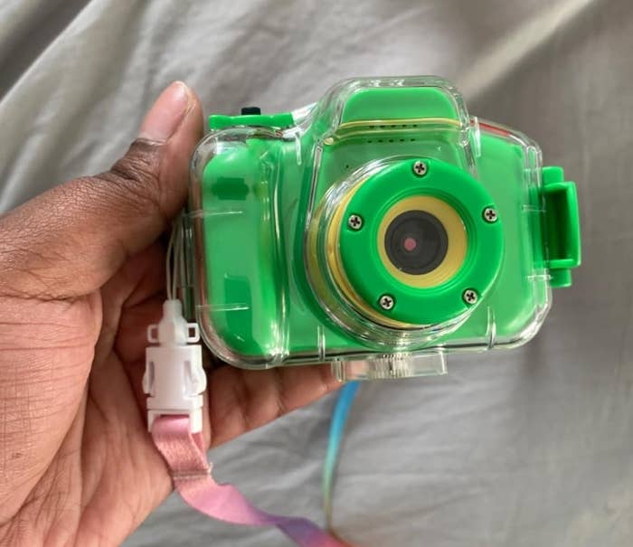 A hand is holding a waterproof, green toy camera with a wrist strap. The camera is encased in a clear plastic cover