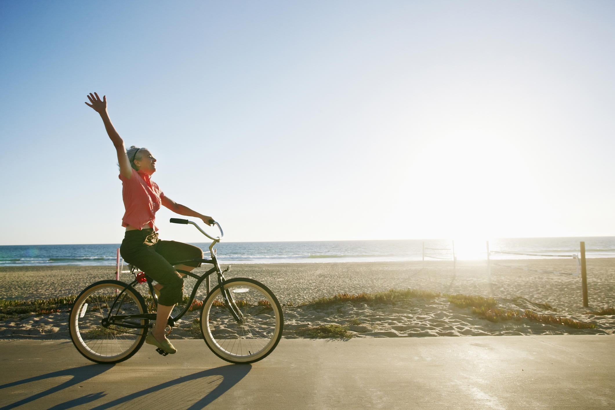 Person riding a bicycle along a beach, one arm raised in joy as the sun sets on the horizon