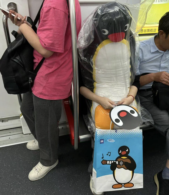 Person sitting on a subway, wearing a Pingu costume covered by a plastic raincoat, and holding a matching bag. Another person stands next to them in casual clothing