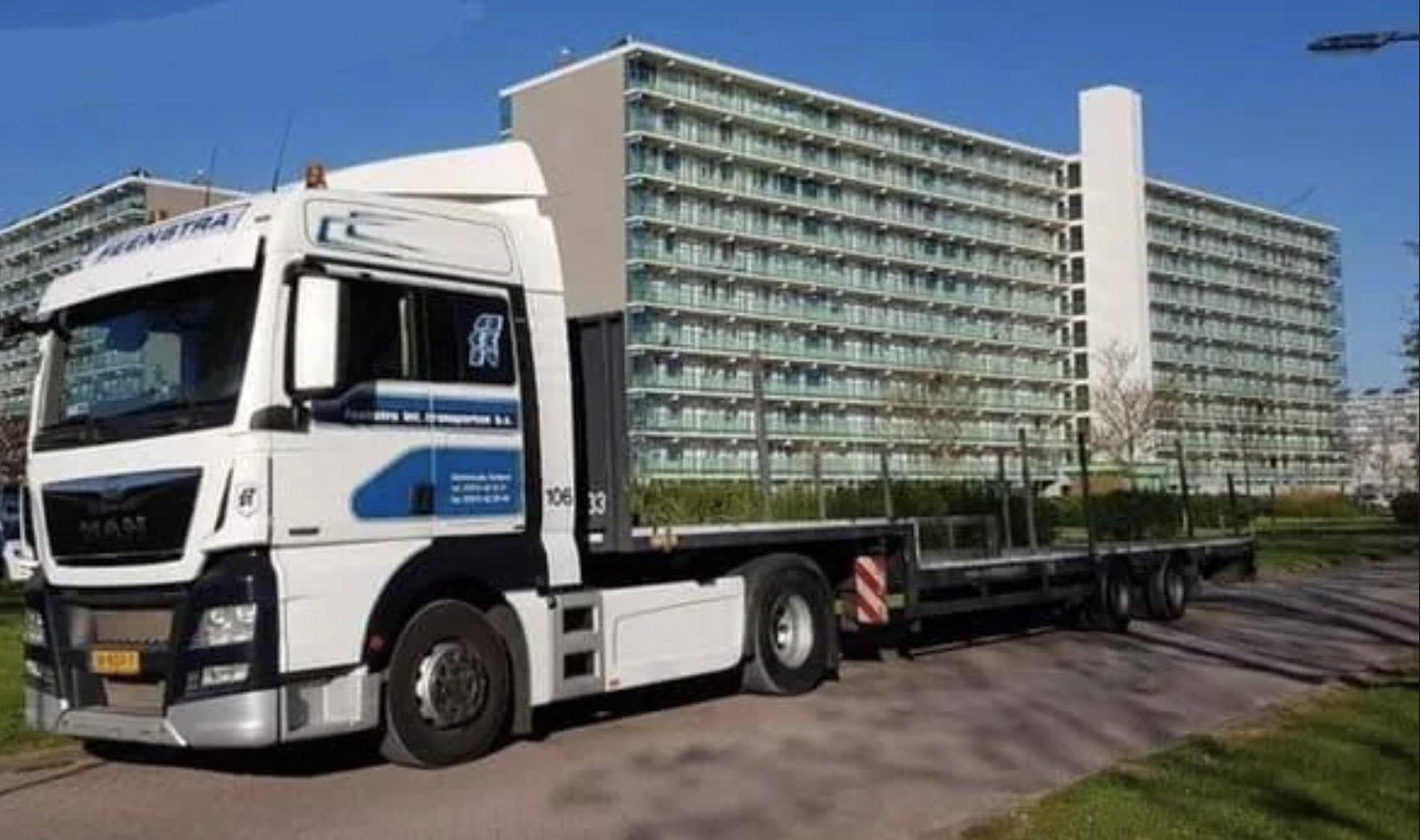 A large truck with a flatbed trailer parked in a residential area, with an apartment building in the background