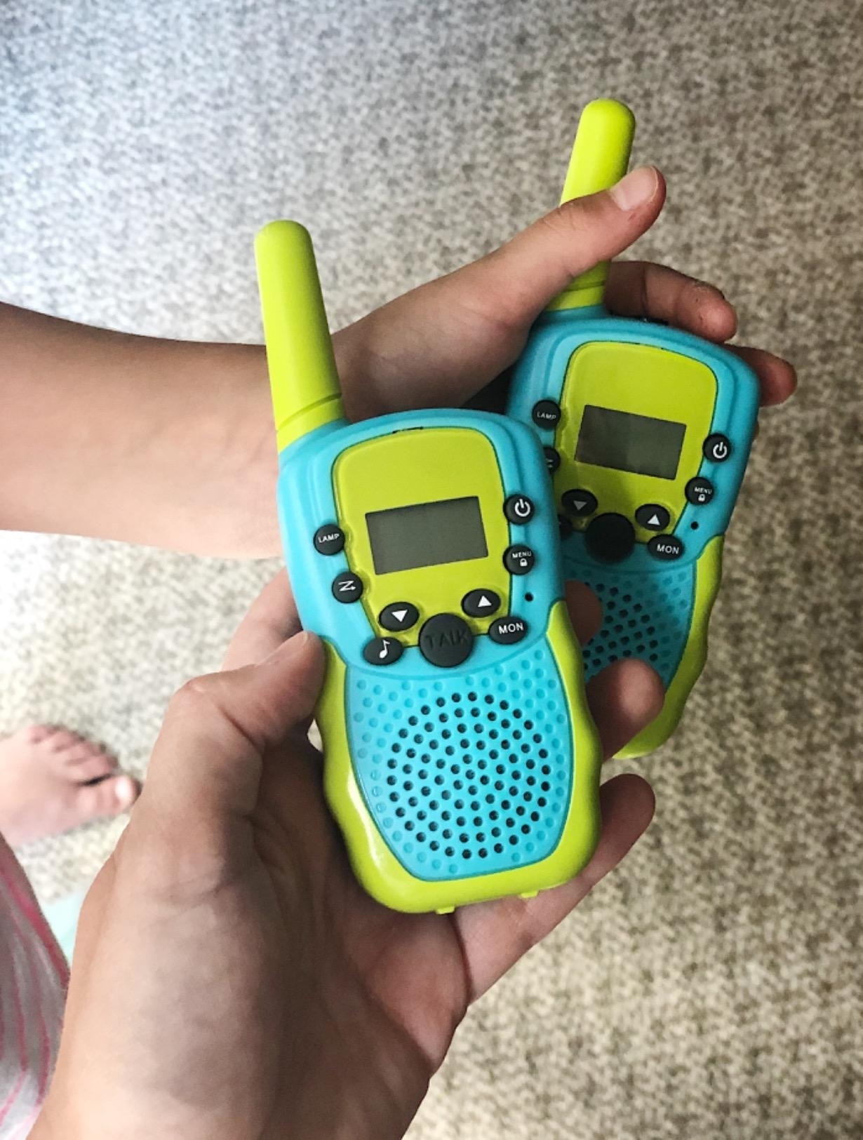 Two hands holding a pair of blue and green walkie-talkies with buttons and a small screen, suitable for kids&#x27; toys