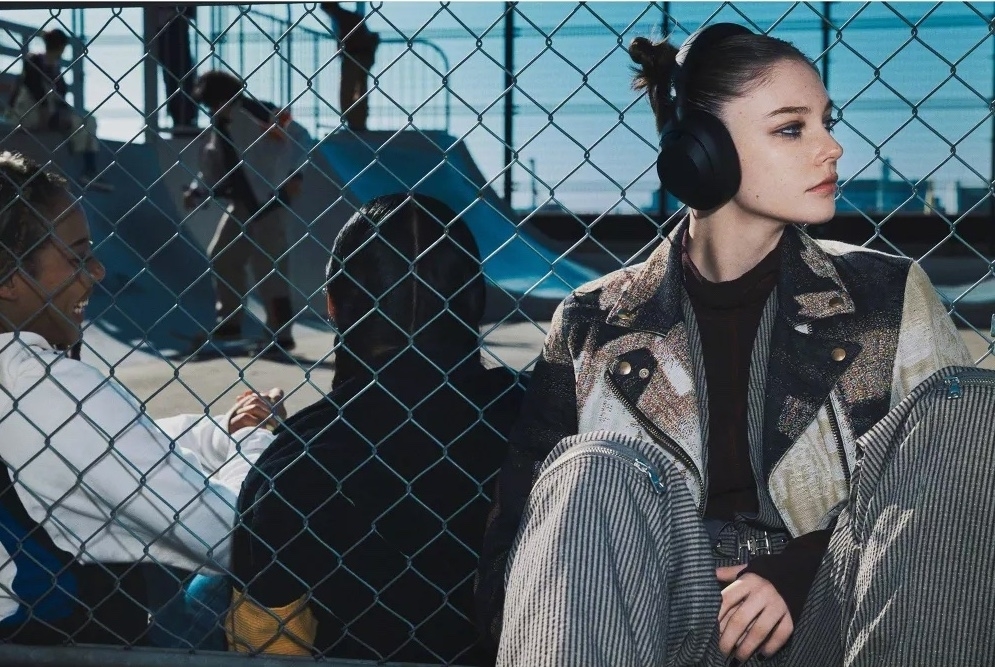 Model in striped pants and patchwork jacket with headphones poses by a fenced outdoor skate park. Two skateboarding friends sit and interact in the background