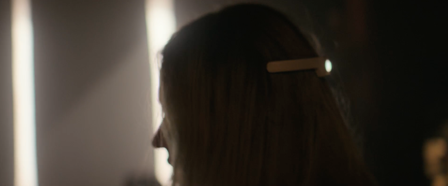 A person with long hair is seen from the side in a dimly lit setting, with a clip visible in their hair. The background is out of focus