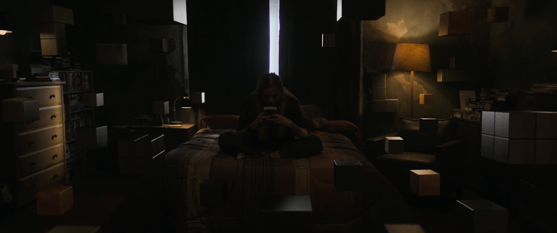 A person sits on a bed in a dimly-lit room with floating cubes surrounding them, concentrating on a handheld device. There is a lamp on the bedside table