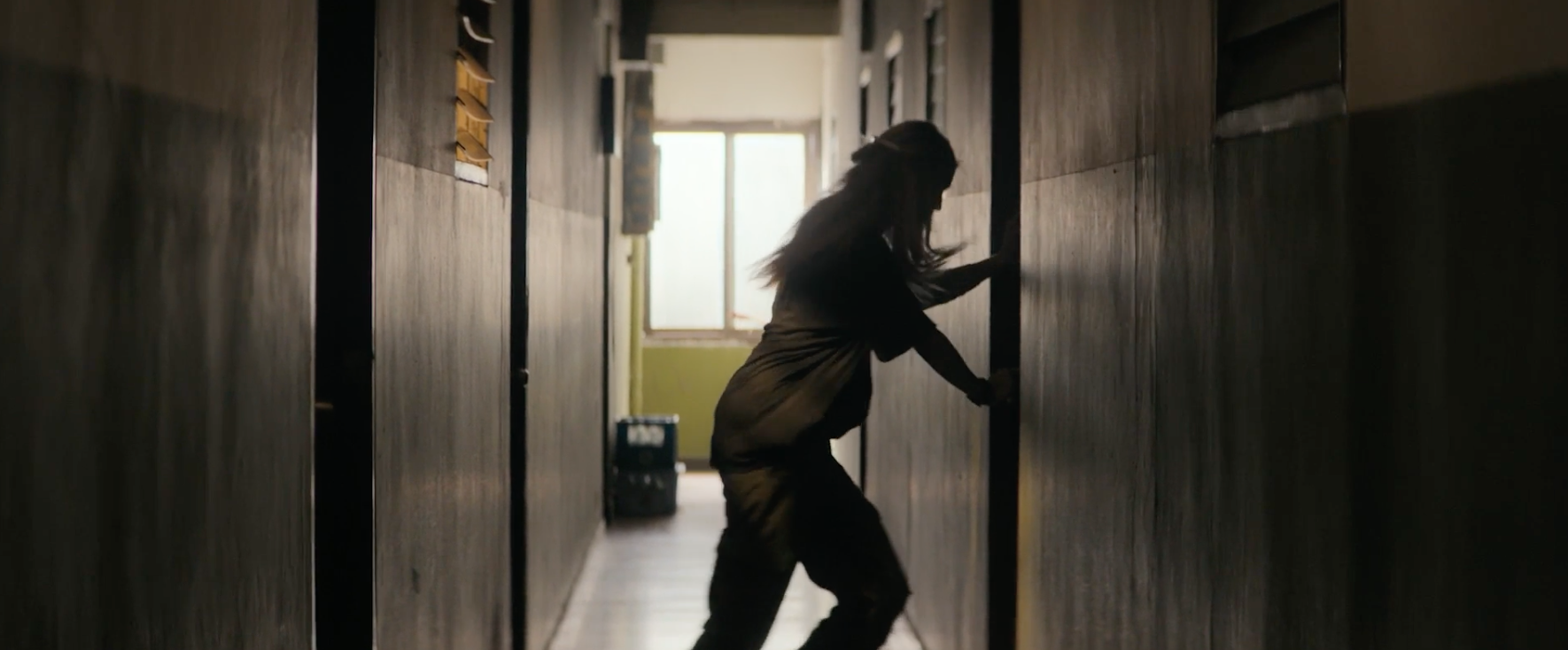 A person in a hallway pushes a door open with effort, silhouetted against a bright window at the end of the hallway