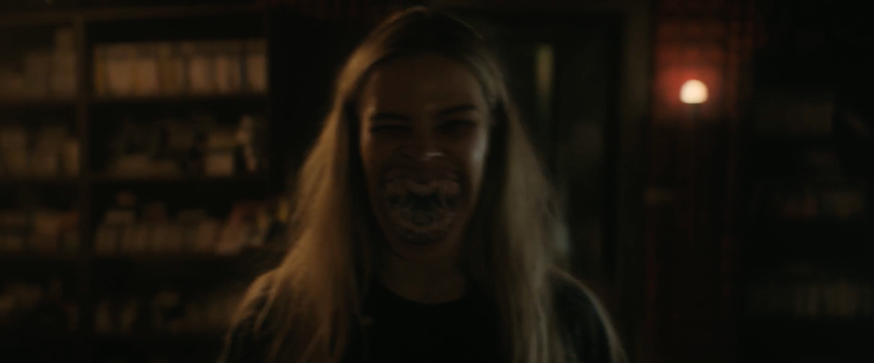 A person with long hair is standing in a dimly lit room with distorted facial features, giving an eerie and unsettling expression