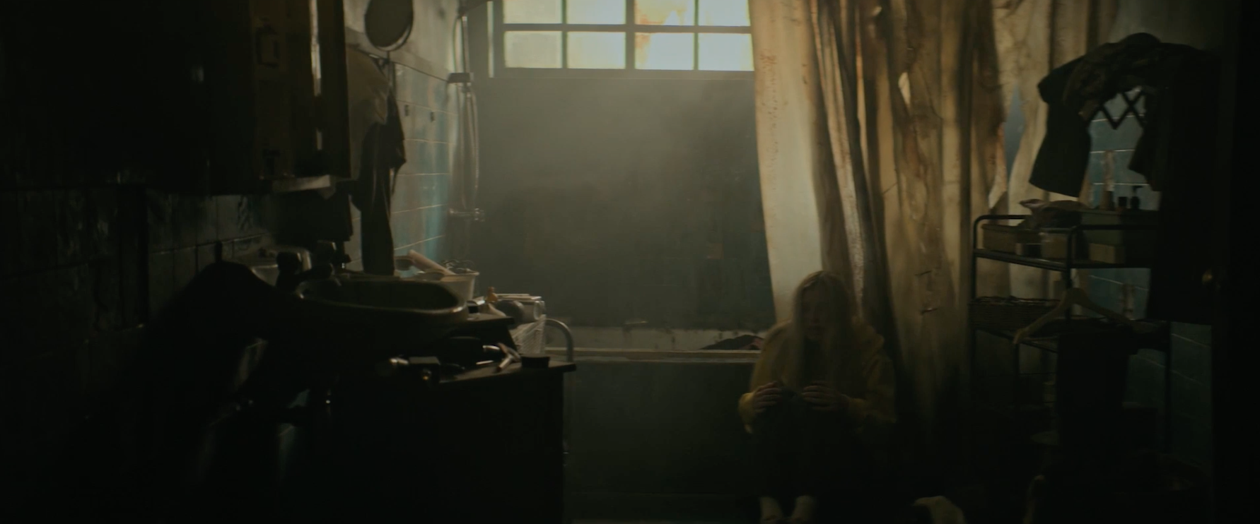 A person sits on the floor in a dimly lit, cluttered bathroom with a bathtub, partially covered by a semi-transparent curtain