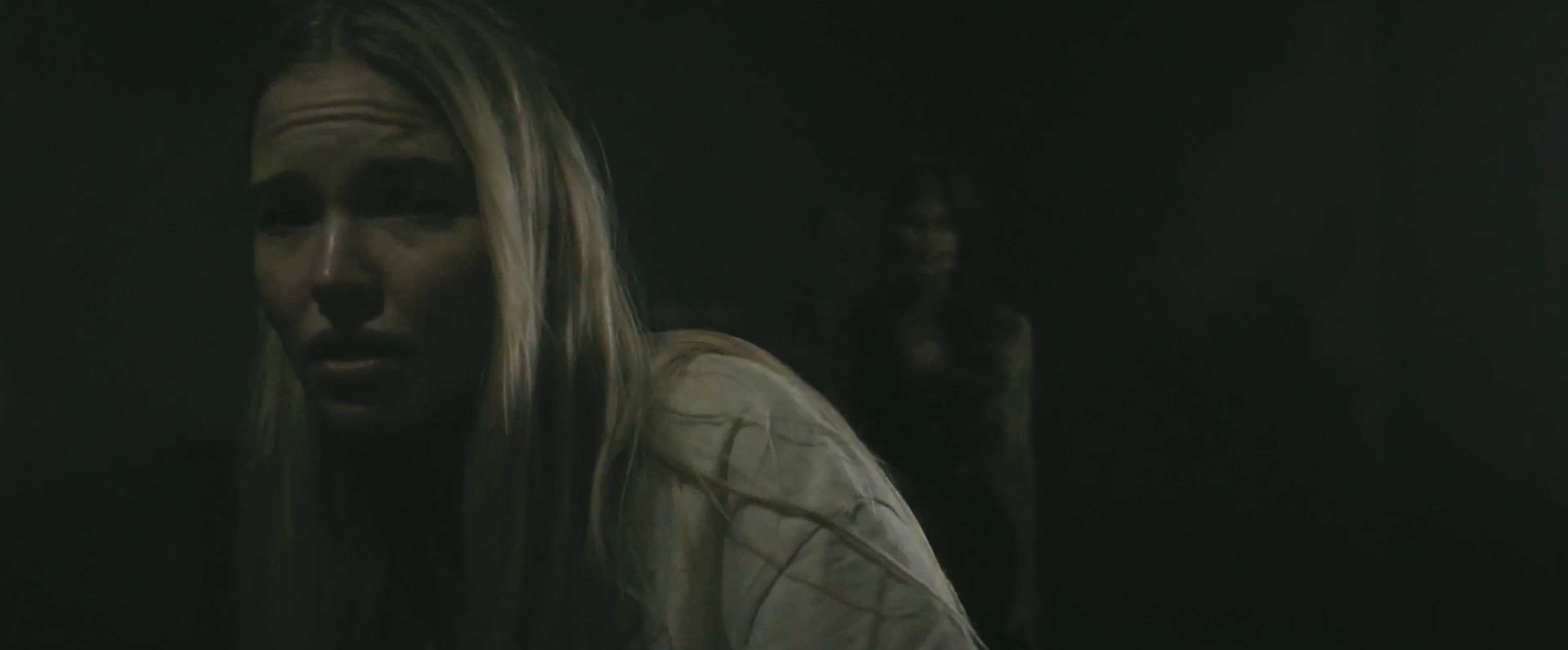 A woman with long, straight hair looks scared, leaning away from a shadowy figure in the background