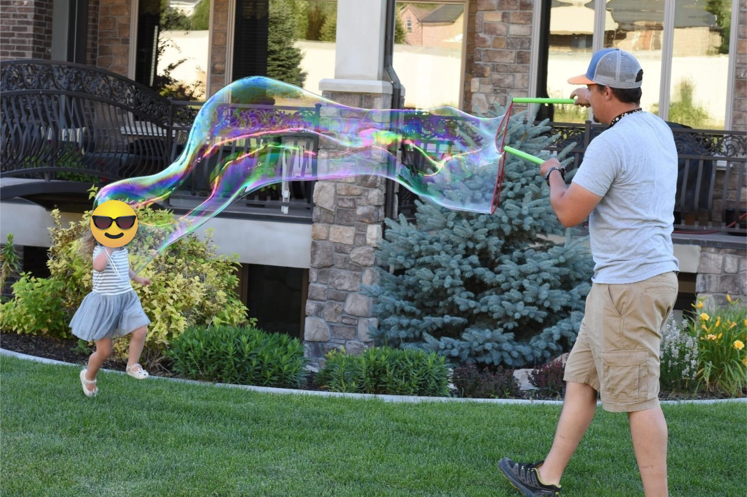 An adult wearing casual clothing creates large bubbles in a garden, while a young child in a striped shirt and skirt runs towards them joyfully