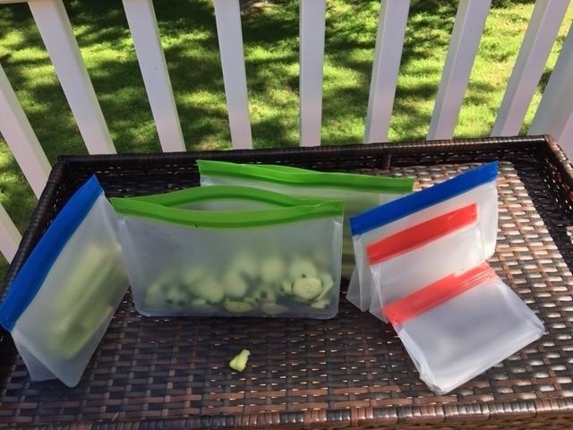 Reusable silicone storage bags are displayed on a wicker tray, some containing chopped vegetables. The bags vary in size and have colored zip closures