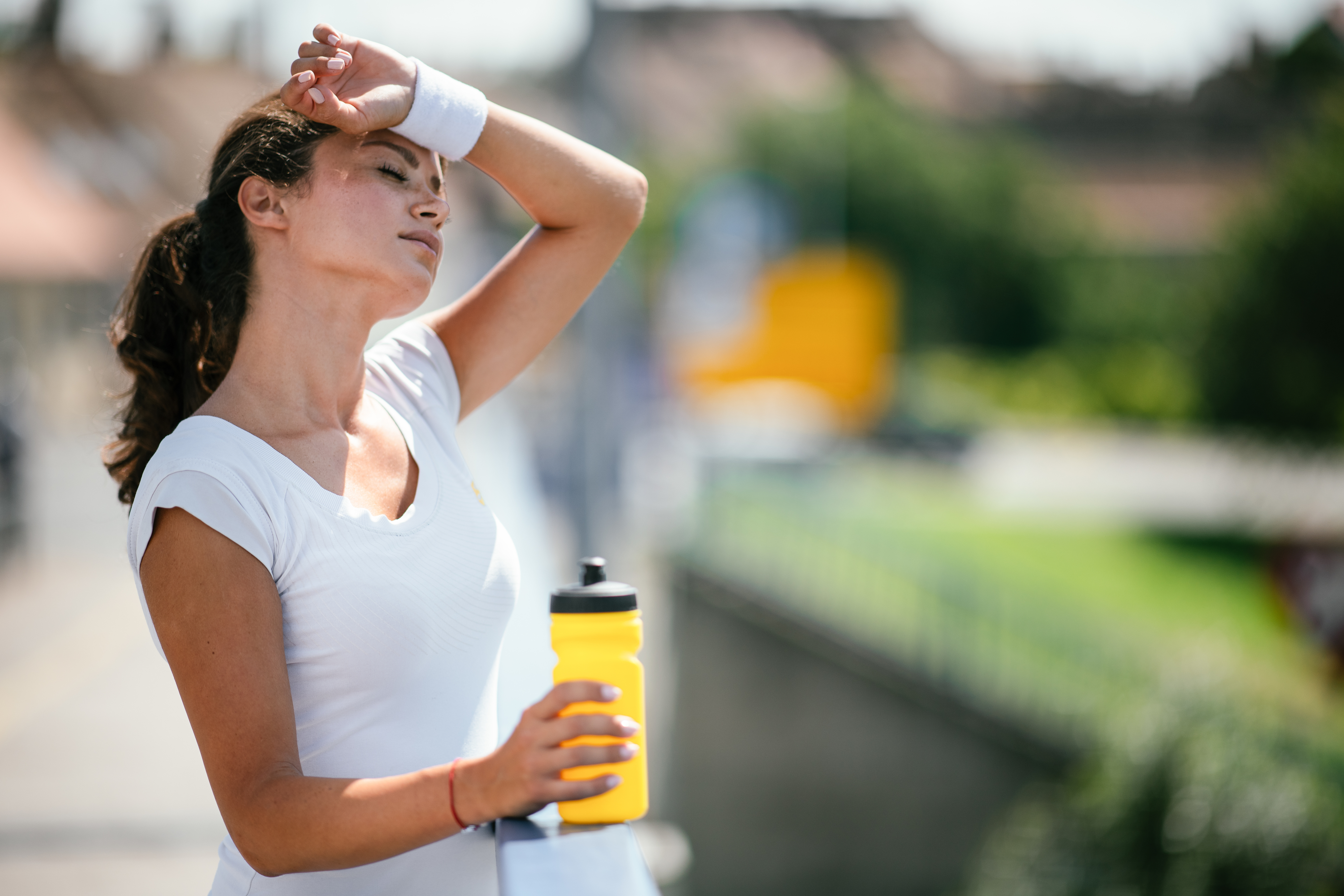 A woman wearing a white athletic outfit and a wristband holds a yellow water bottle, wiping sweat from her forehead, appearing exhausted after exercising outdoors