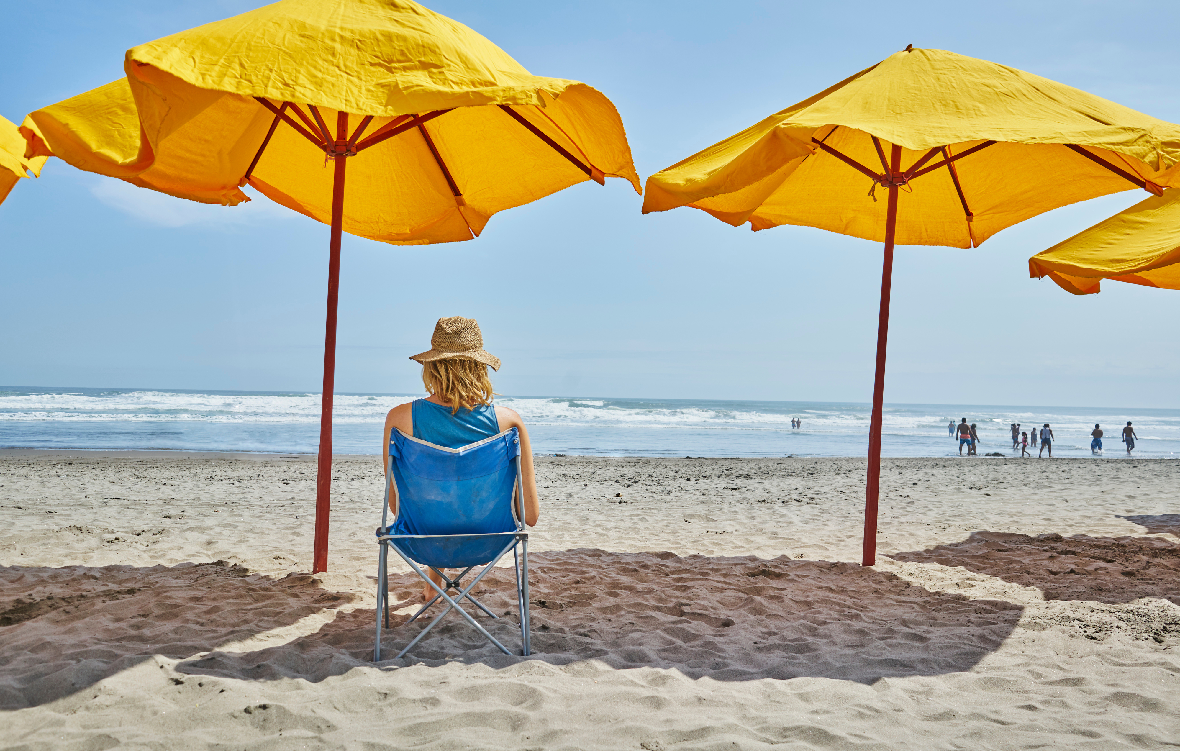 A person in a sun hat sits on a beach chair under yellow umbrellas facing the ocean. Several people can be seen walking along the shoreline in the distance