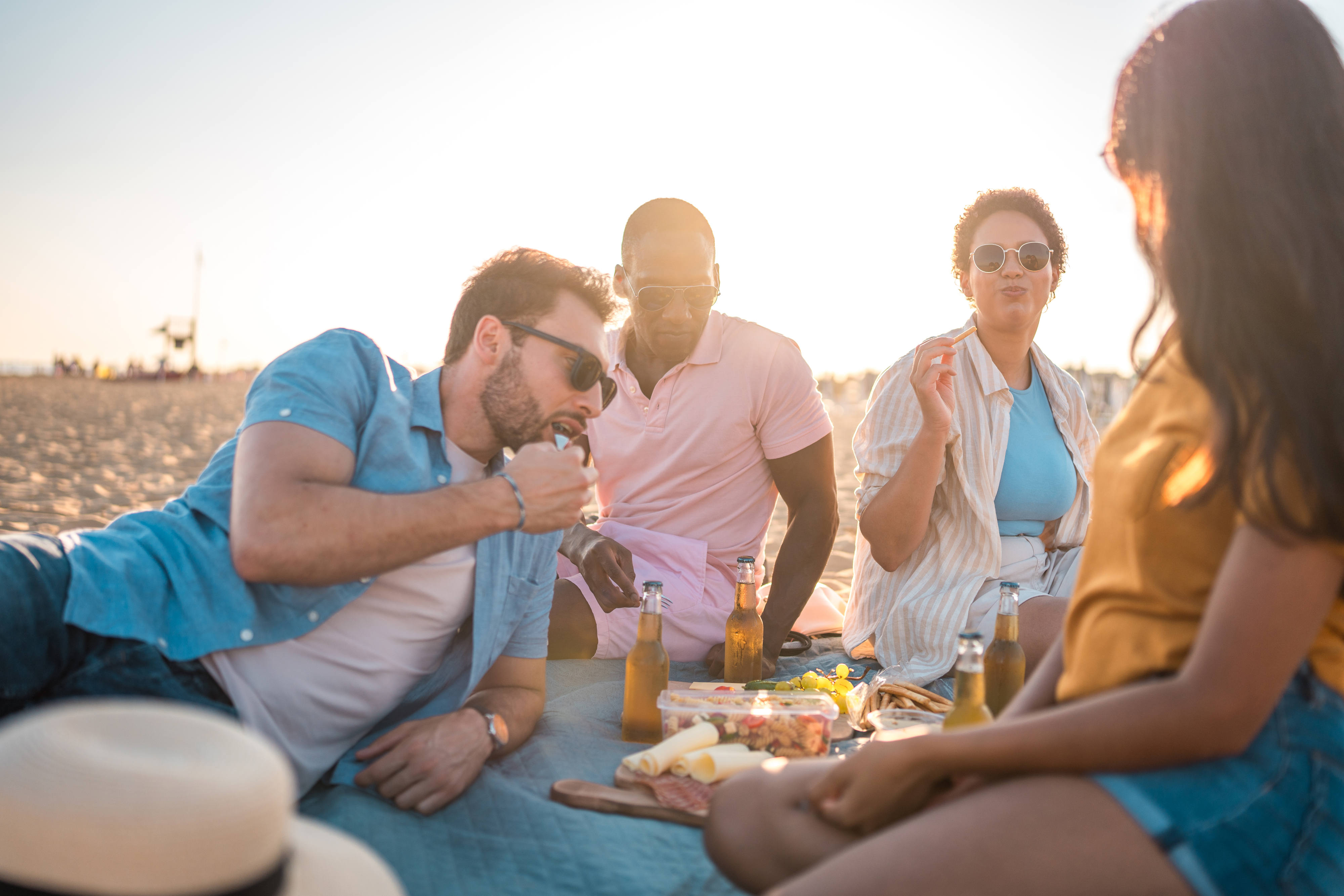 A group of four friends enjoying a picnic on the beach, sharing snacks and drinks, with a warm sunlit backdrop. Names unknown