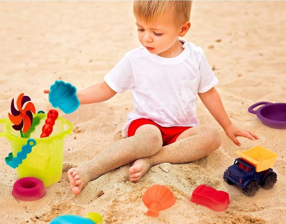 Child in a white shirt and red shorts plays on the sandy beach with colorful toys, including a green bucket, toy seashells, and a toy truck