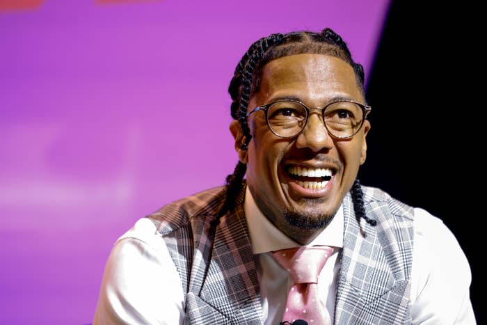 Nick Cannon smiling, wearing glasses, a plaid vest over a white dress shirt, and a pink tie