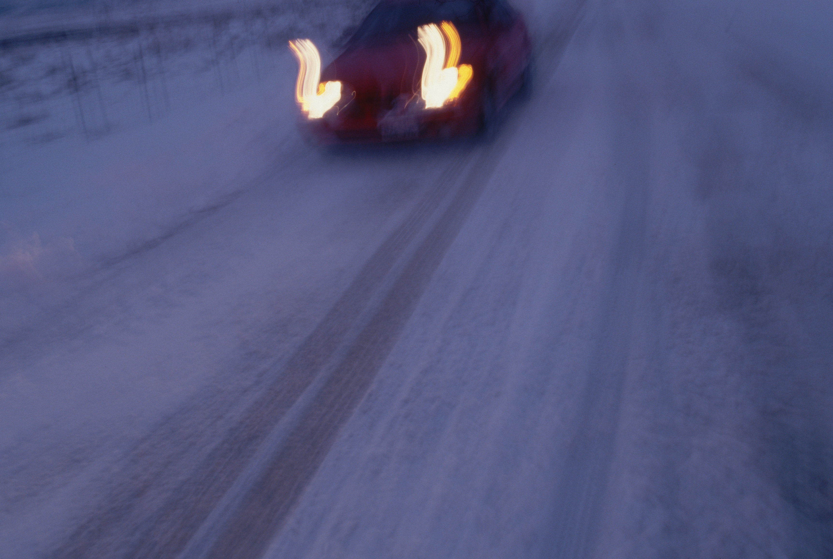 A car driving on a snow-covered road at night with headlights on, creating a blurred effect. No identifiable persons are in the image