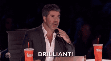 Simon Cowell wearing a suit and sitting at a table with two drink cups, gesturing and saying &quot;Brilliant!&quot; on America&#x27;s Got Talent