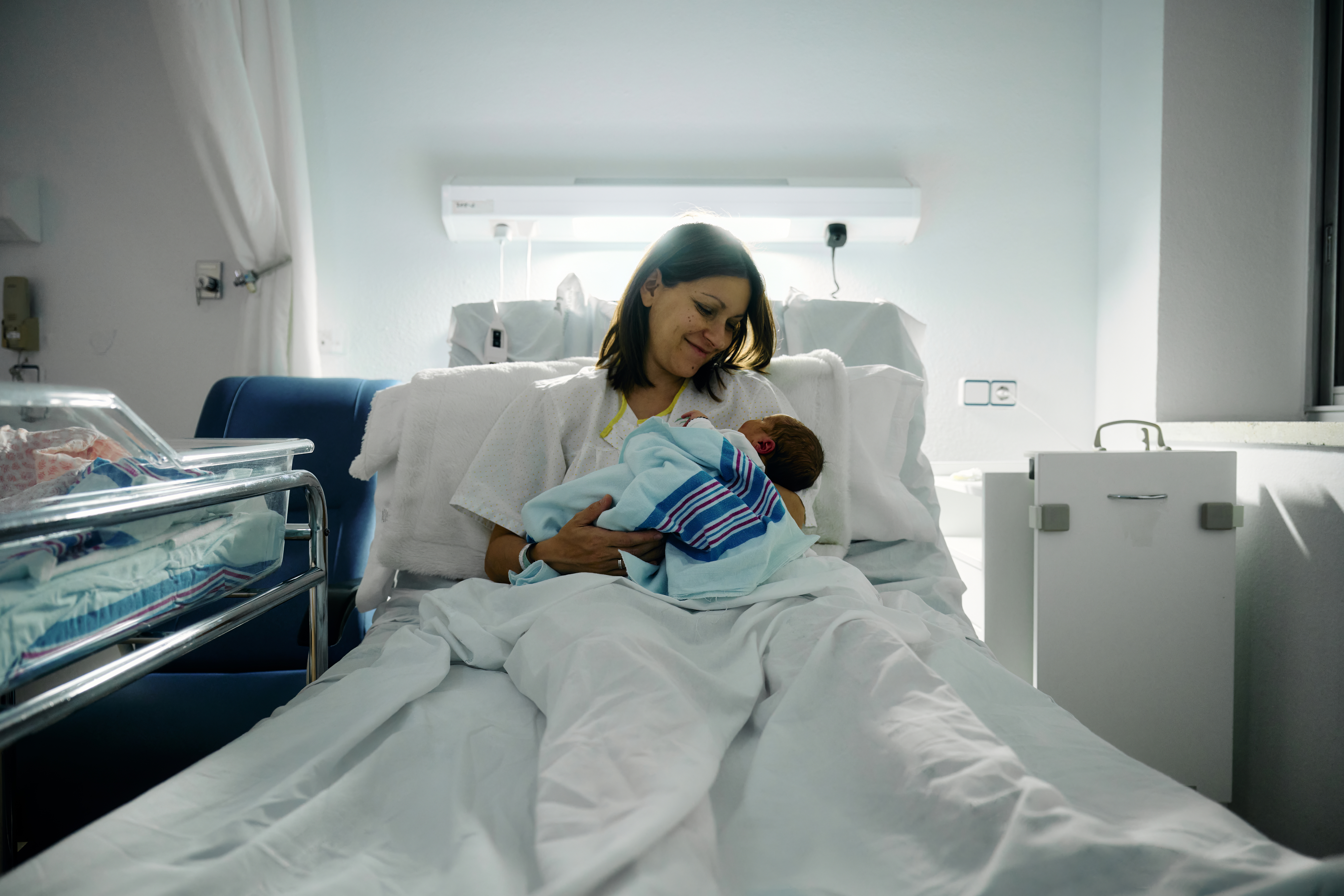 Woman in hospital bed holding a newborn baby wrapped in a blanket, looking down and smiling. A bassinet is seen beside the bed