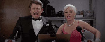 Elizabeth Banks and John Michael Higgins dressed formally, reacting animatedly while seated at a desk with microphones