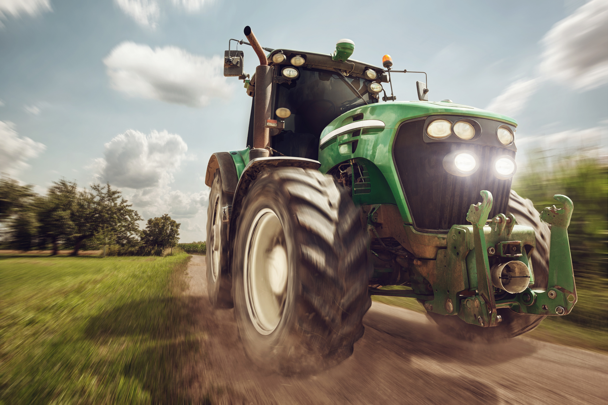 A green tractor drives down a rural dirt road with grassy fields and trees in the background under a partly cloudy sky