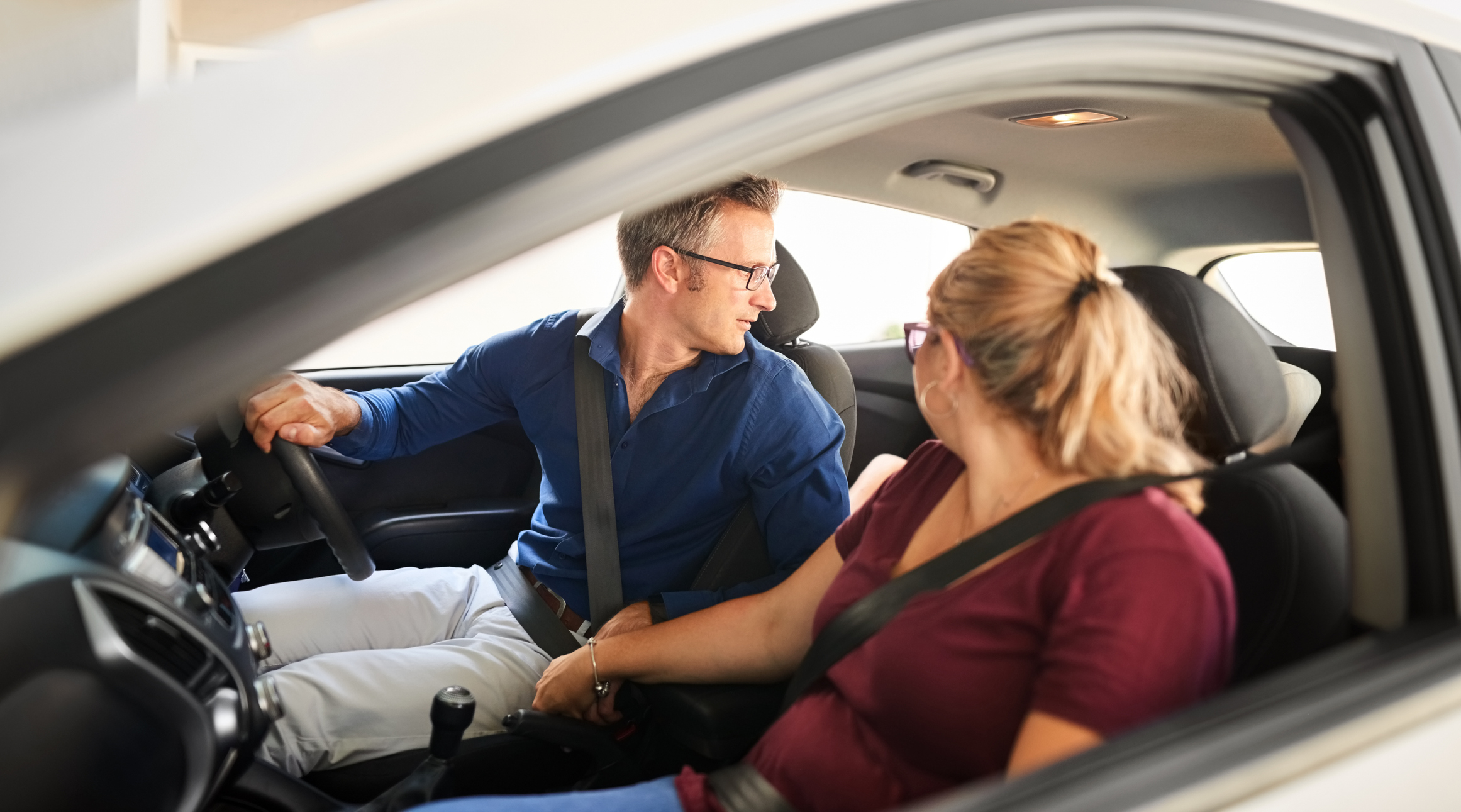 A man and a woman sitting in a car. The man is in the driver&#x27;s seat looking back, while the woman in the passenger seat looks at him