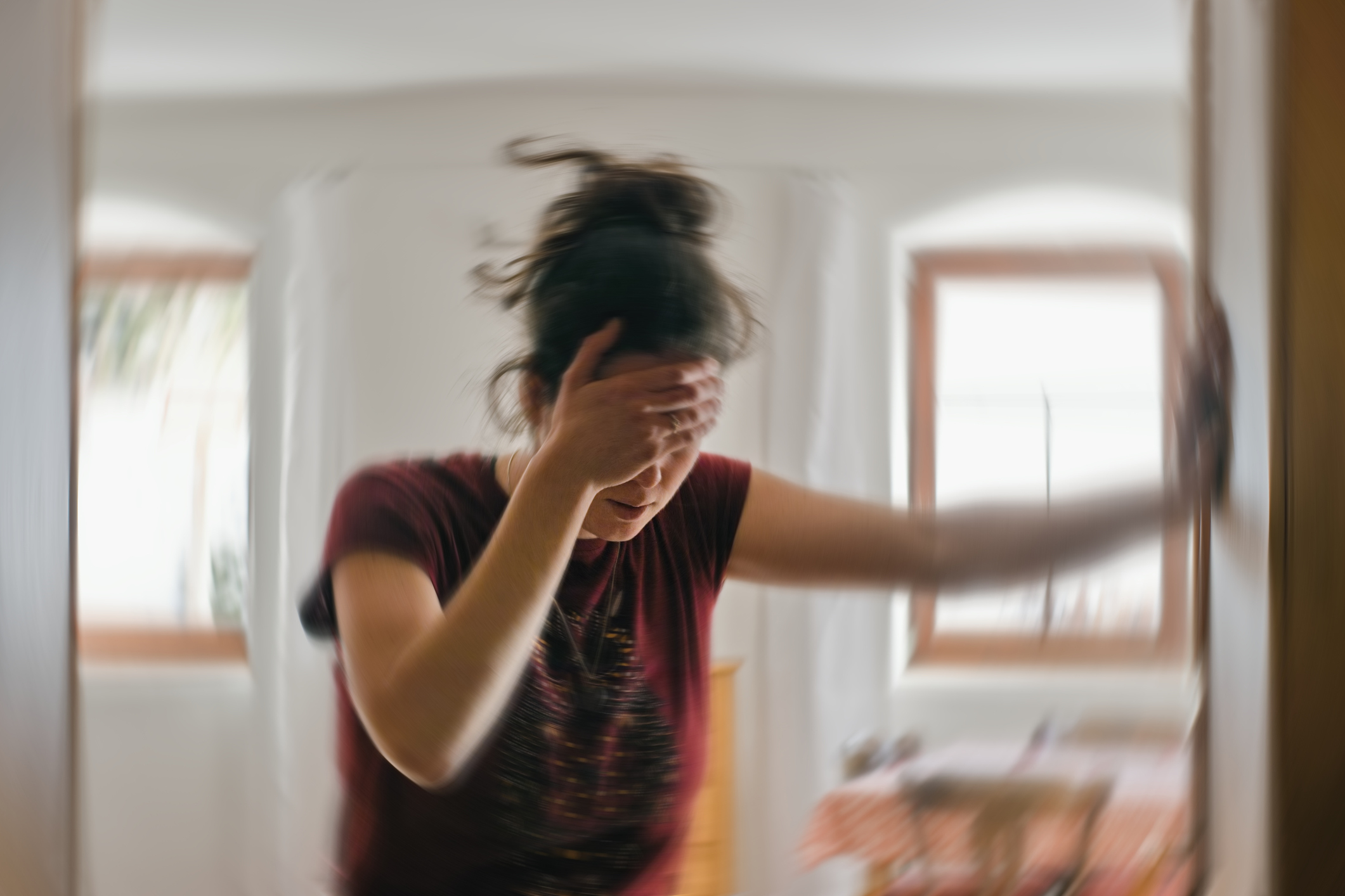 A person holding their head in pain, standing in a blurred room, looking distressed