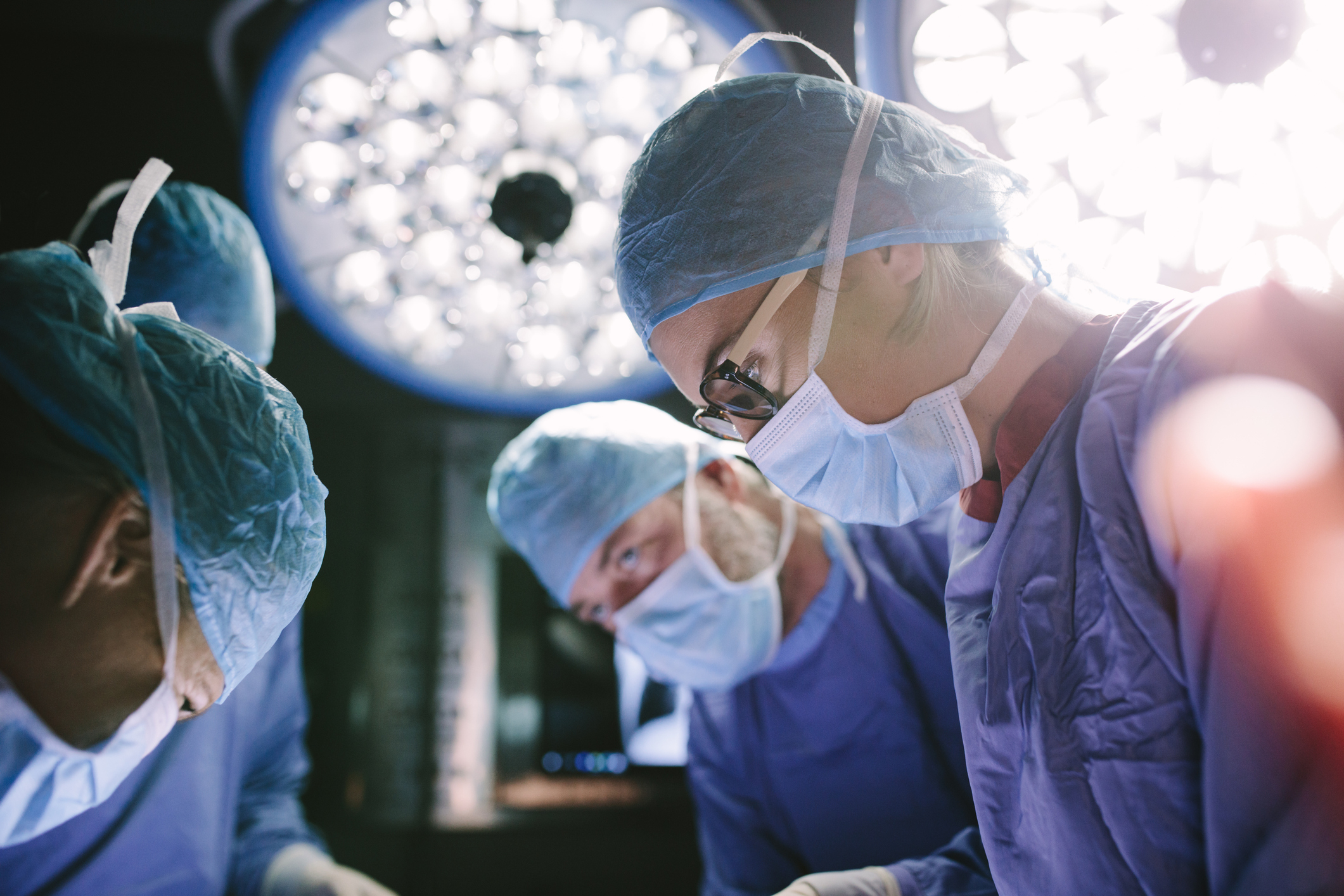 Surgeons perform a procedure in an operating room, wearing scrubs, masks, and caps, with surgical lights overhead