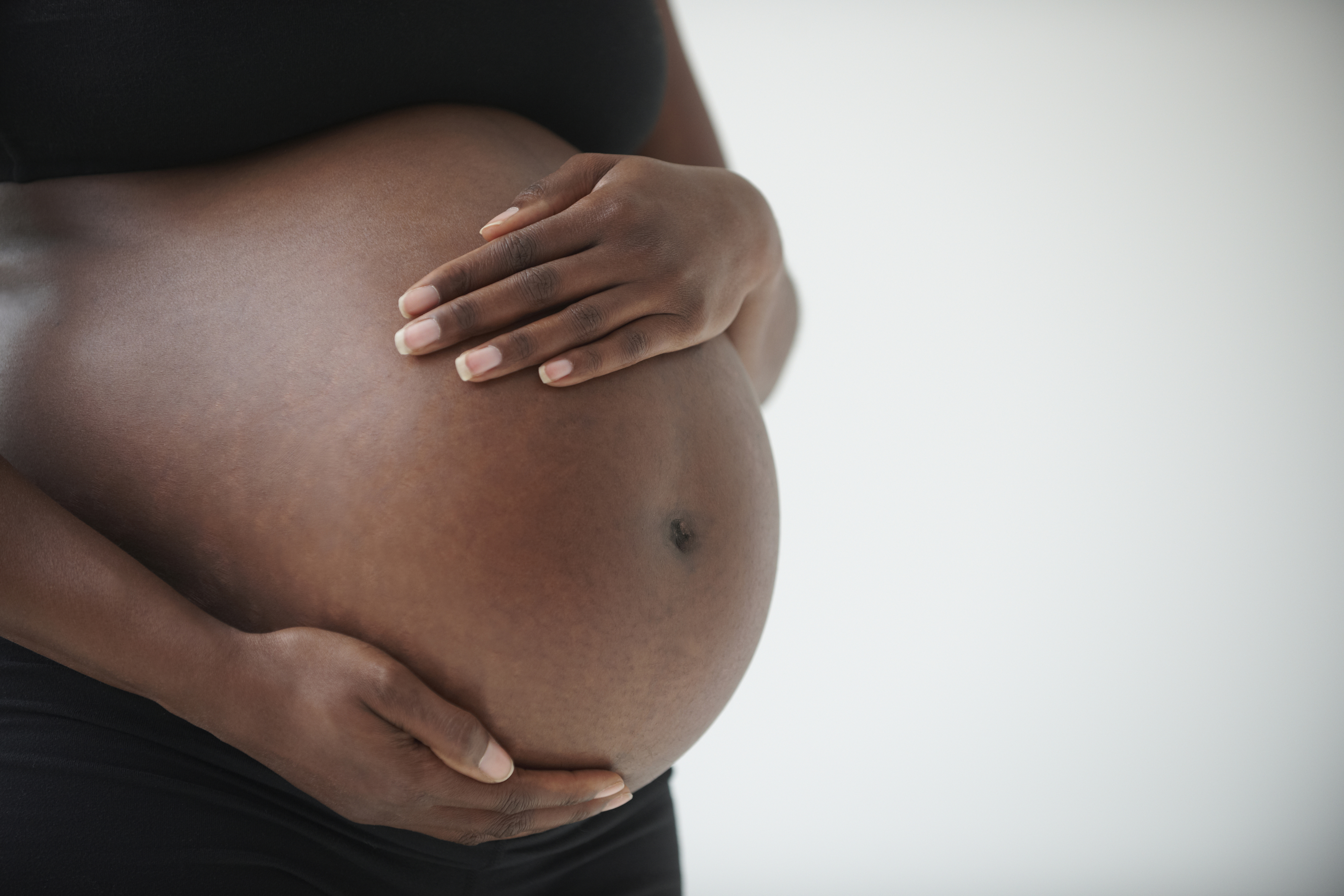 A close-up of a pregnant woman&#x27;s belly with her hands gently resting on it. The woman is wearing a black top and pants