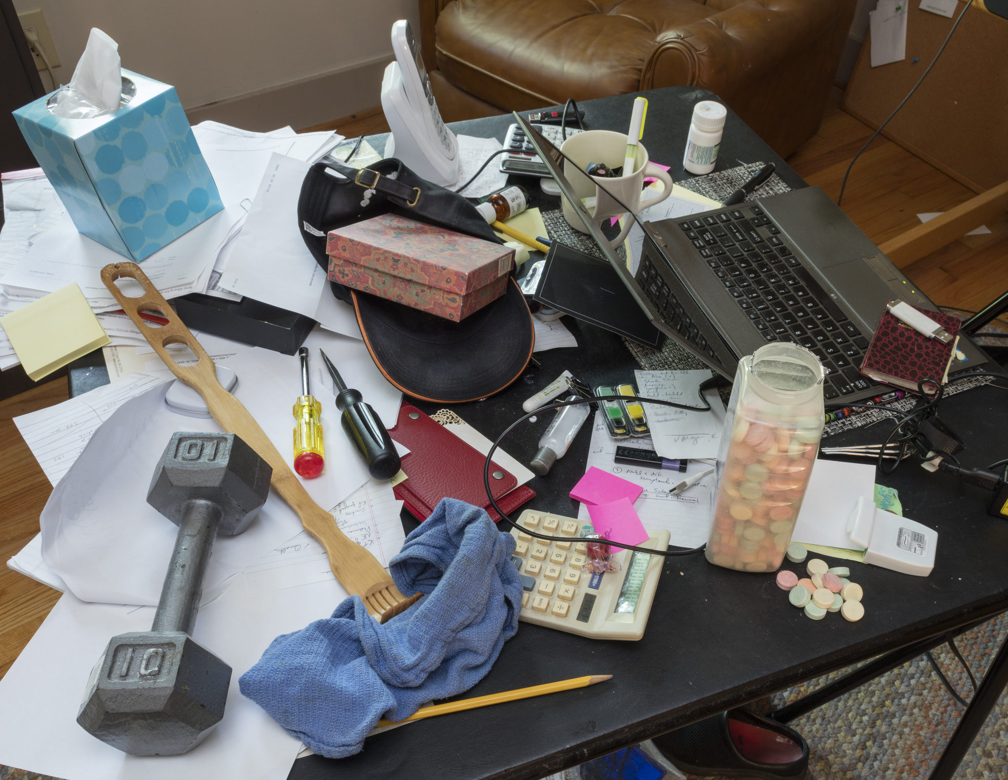 A cluttered desk includes a laptop, tissues, dumbbell, calculator, various papers, a screwdriver, and a container of candy