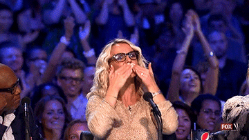 A woman wearing glasses and a sparkly outfit blows kisses to the audience while seated at a panel with two others