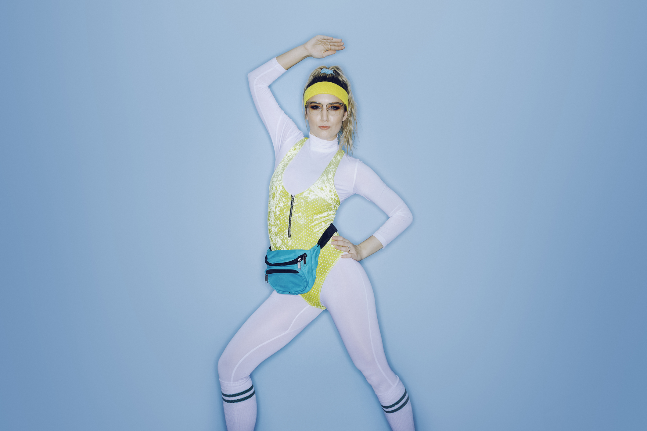 Woman in a retro workout outfit posing confidently. She&#x27;s wearing a yellow headband, blue fanny pack, white leotard, and yellow lace unitard. Her arm is raised