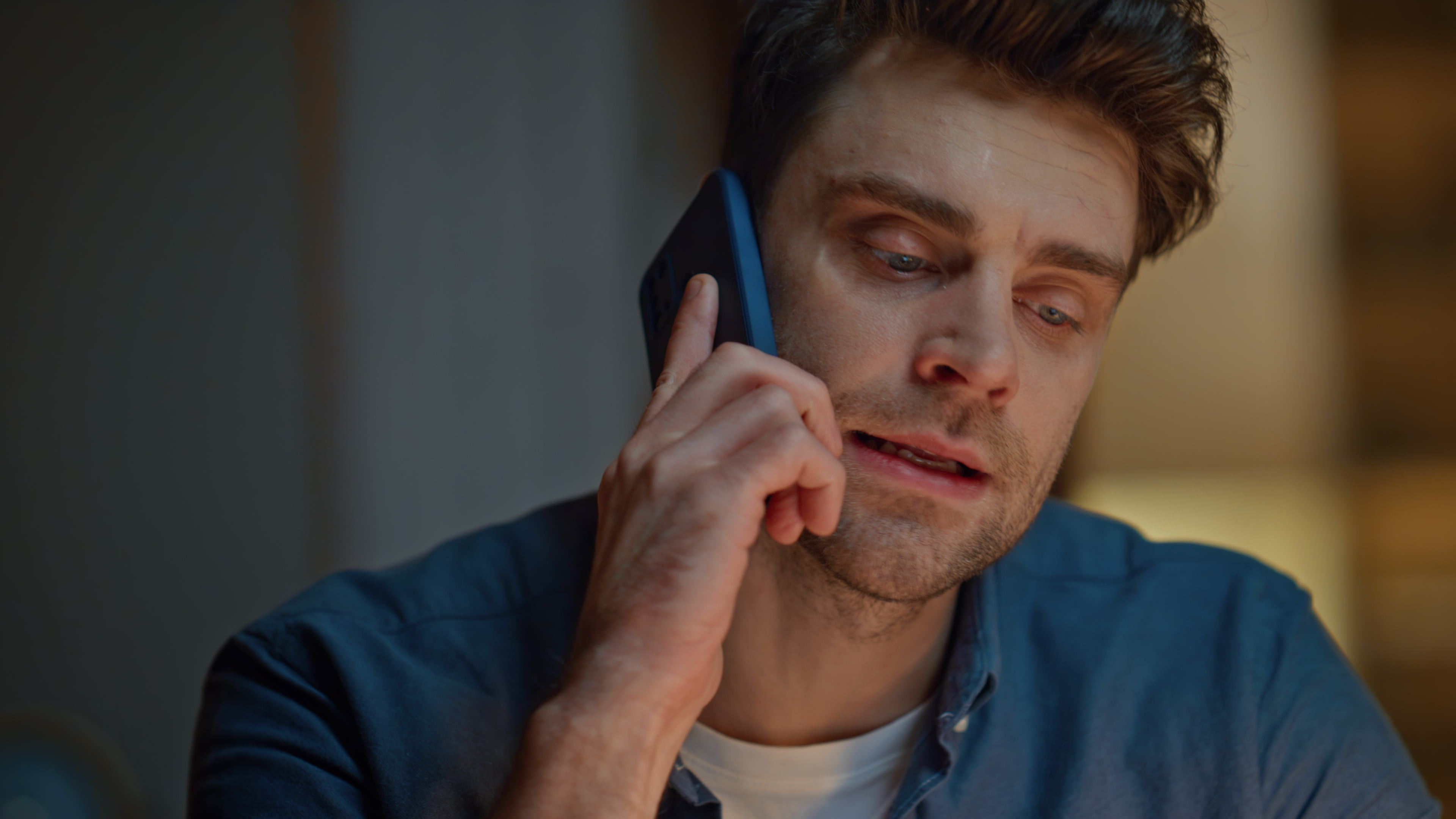 A man with short hair and beard is on the phone, looking down pensively while wearing a shirt