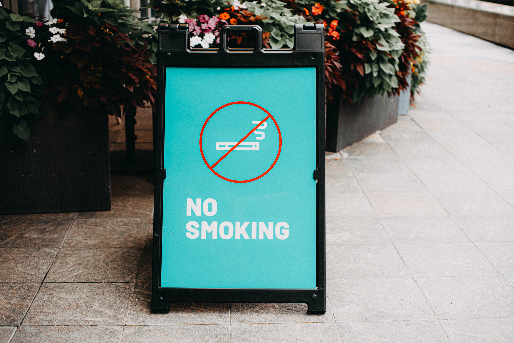 A sidewalk sign with the message &quot;No Smoking&quot; and an icon of a cigarette crossed out, placed next to planters with flowers