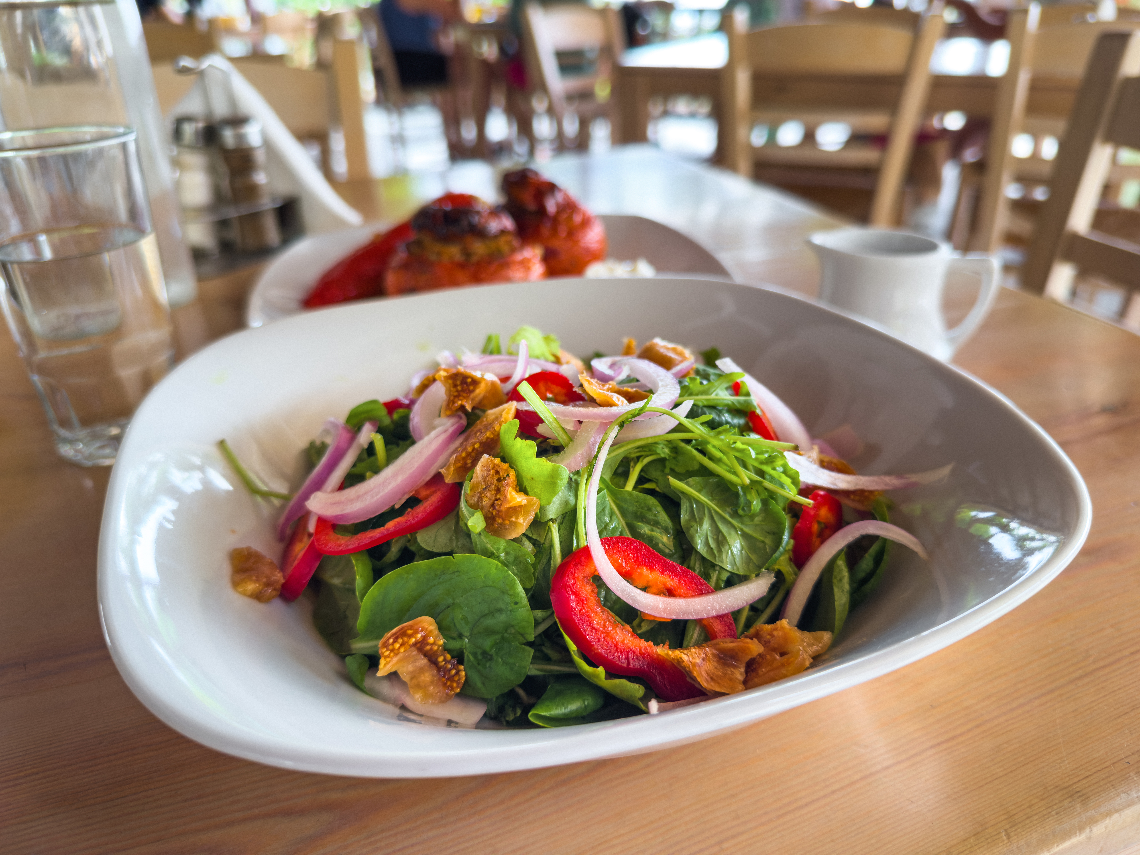 A fresh salad with leafy greens, red bell pepper strips, thinly sliced onions, and fig pieces in a white bowl, set on a wooden table in a restaurant
