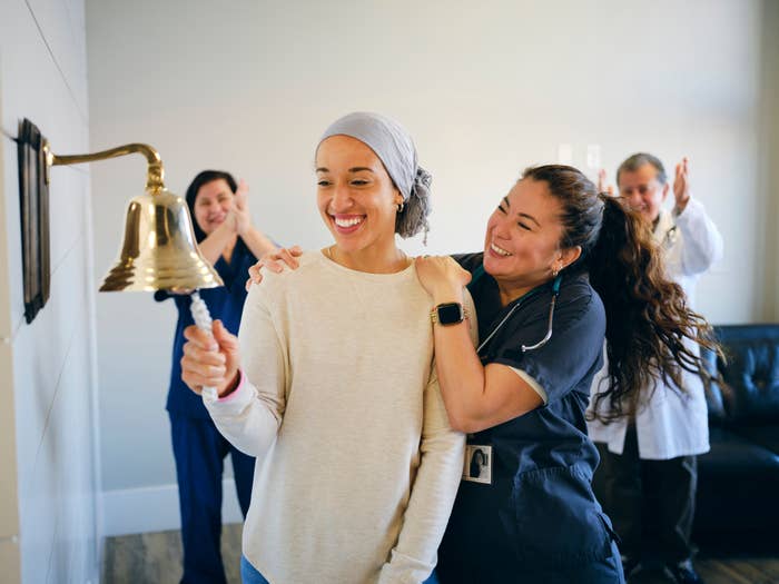 A woman wearing a headscarf rings a bell while a nurse and two doctors applaud, celebrating the end of her treatment