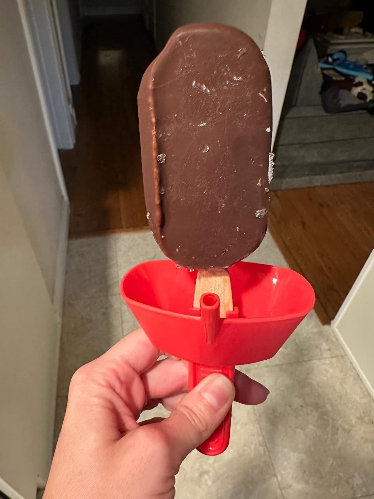 A hand holding a chocolate-covered ice cream bar with a red drip tray attached to the stick