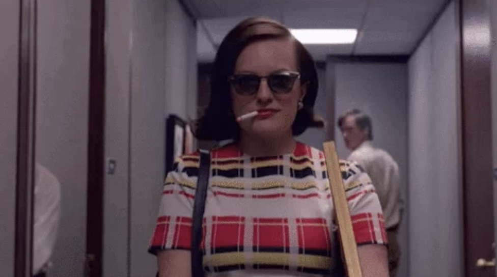 Elisabeth Moss walking down a hallway, wearing sunglasses and holding a cigarette, dressed in a patterned dress. A person is seen walking behind her