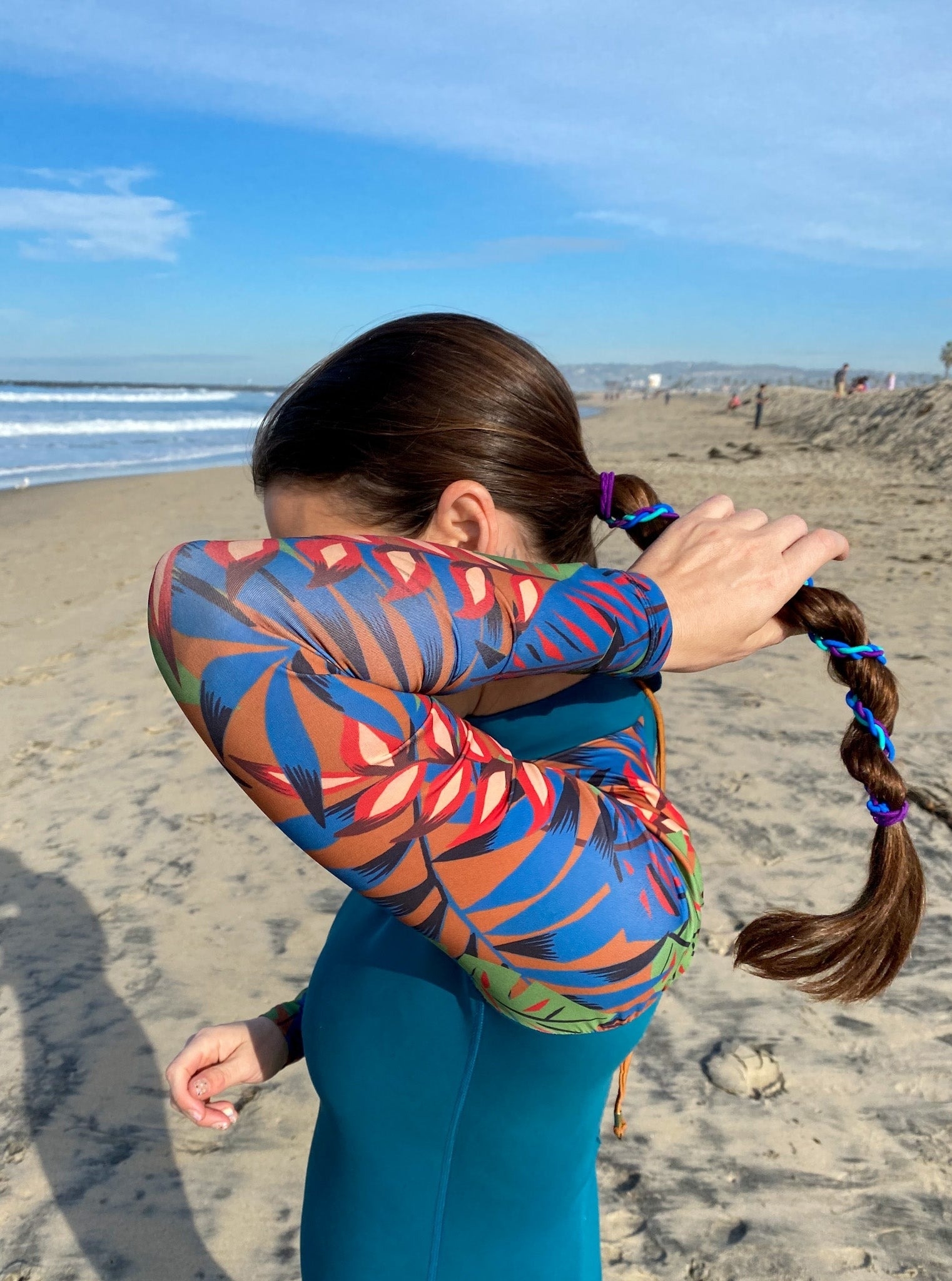 Person on a beach pulling their braided hair, wearing a colorful long-sleeved swimsuit with a pattern of leaves and shapes