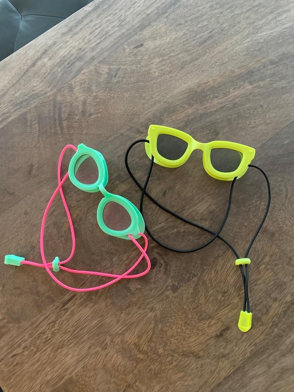 Two pairs of playful sunglasses with attached cords, featuring neon green and pink rims shaped like cat-eye and oval frames, displayed on a wooden table