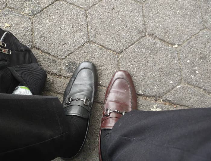 One person wearing mismatched black and brown loafers standing on a hexagonal stone pavement. Black pants are visible