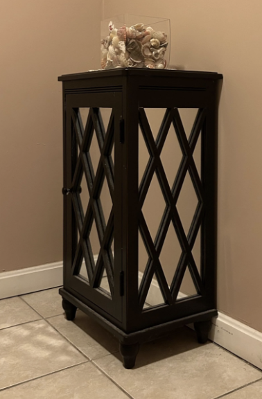 Black cabinet with a glass door and diamond-shaped patterns, placed on a tiled floor. A glass container filled with seashells is on top