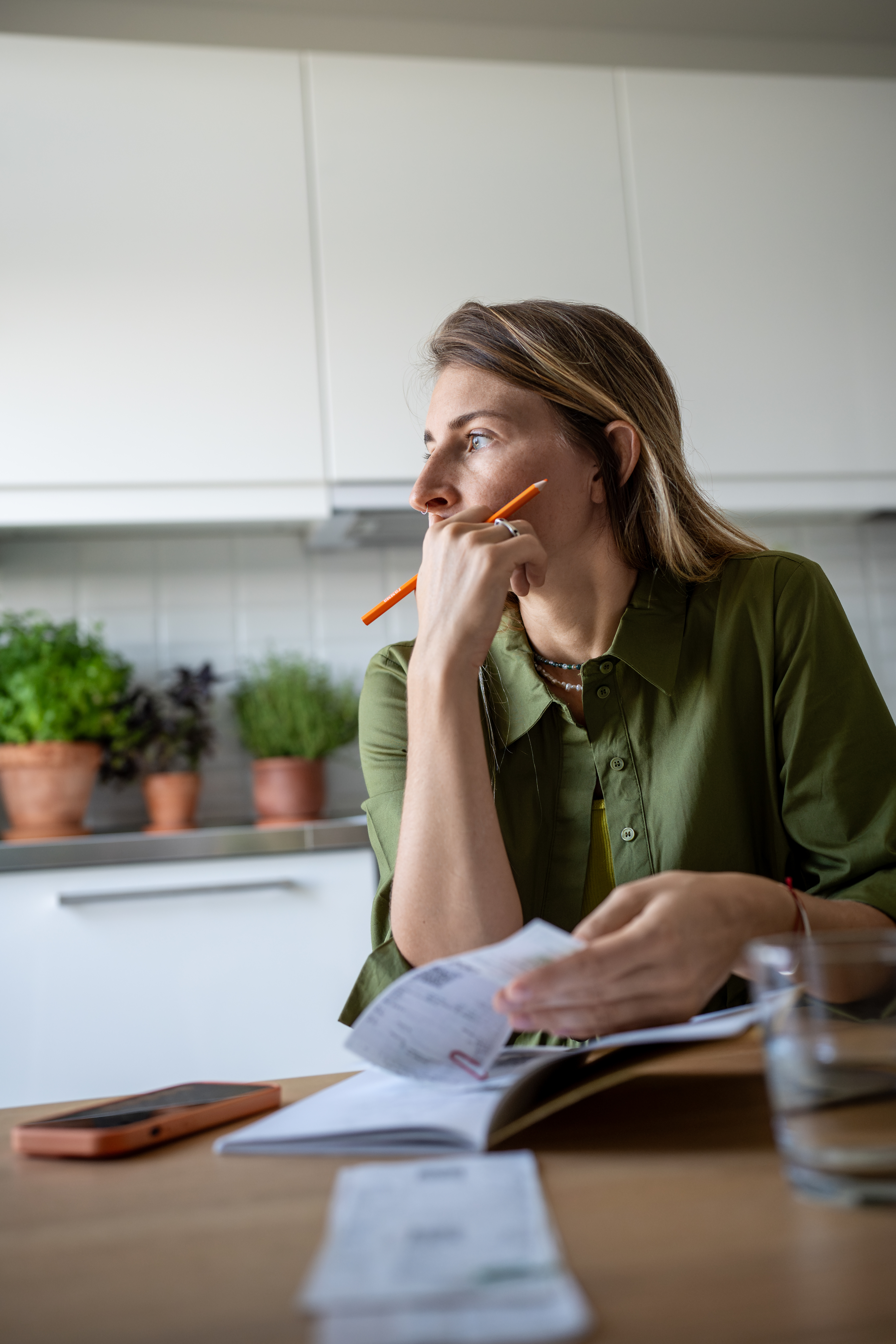 A woman in a green shirt sits at a kitchen table, holding a pencil and looking thoughtful while reviewing papers. Pots with plants are visible in the background