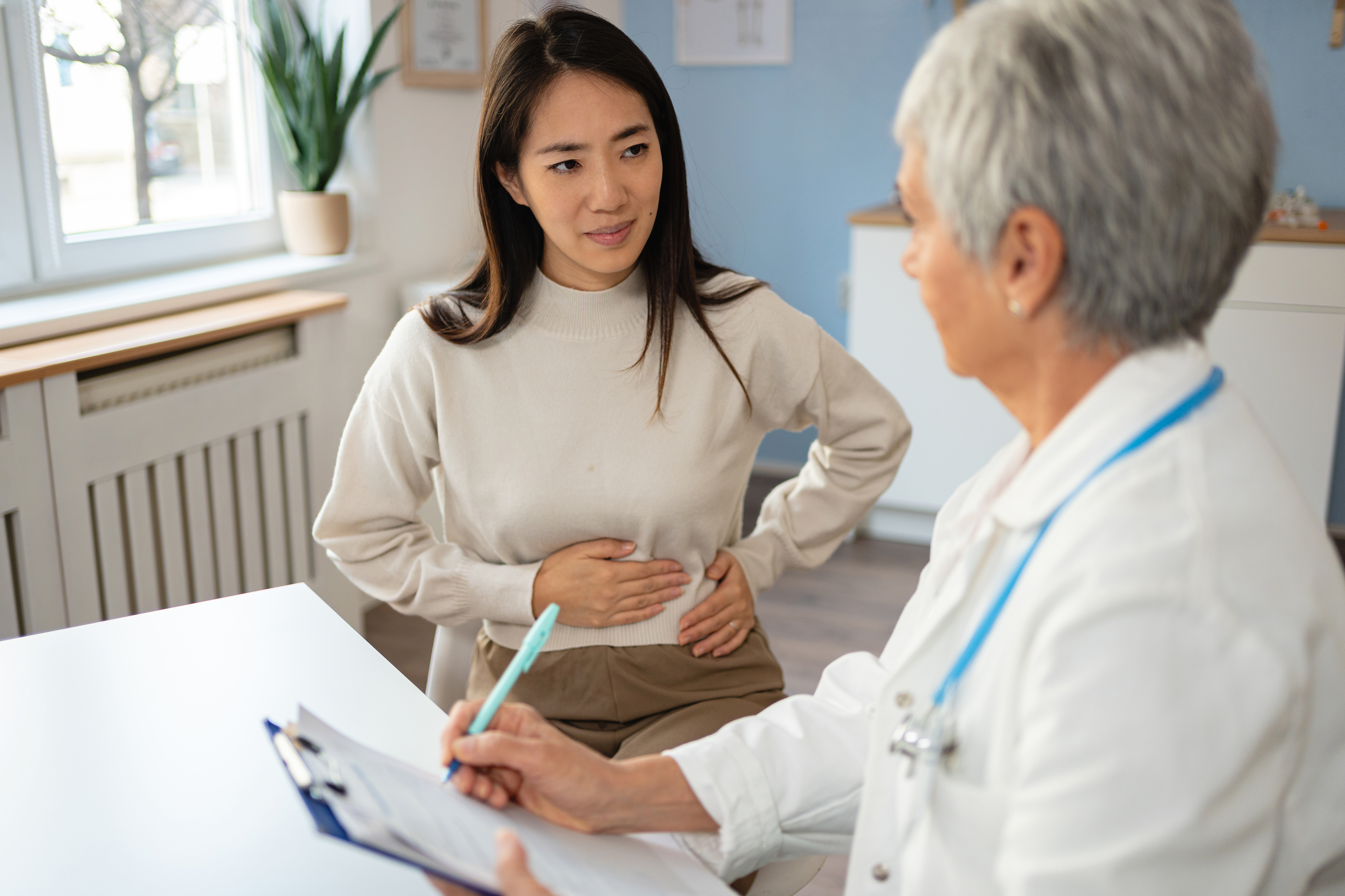 A woman holds her stomach while talking to a doctor in a white coat holding a clipboard